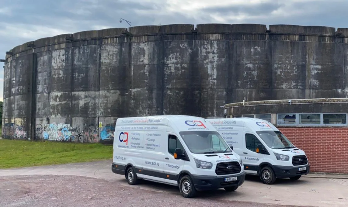 Two AQL white commercial vans parked on a concrete surface in front of a large, weathered cylindrical industrial structure.