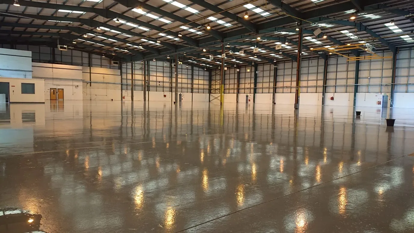 Interior view of a large, empty industrial warehouse with polished concrete floors reflecting overhead lights and a high ceiling supported by metal beams.