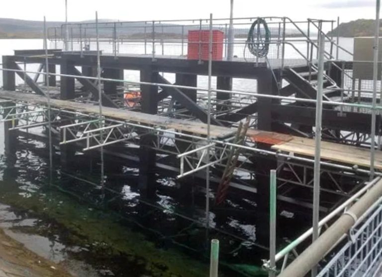 A partially constructed or under-maintenance dock or platform over water, supported by metal scaffolding and beams, with safety railings and a large red container on the upper level.