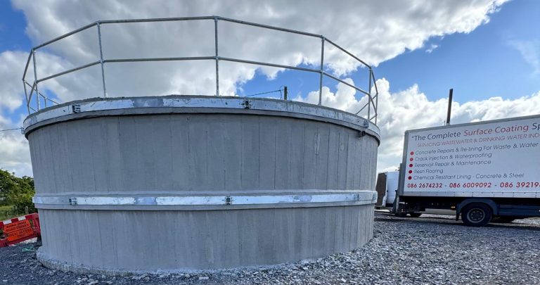 Concrete circular water tank with metal bands and railing on top, set on a gravel surface under a partly cloudy sky, with a white service truck parked nearby.