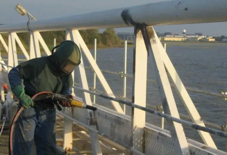 Person wearing protective gear using a heating torch to burn graffiti off a metal railing on a waterfront structure.