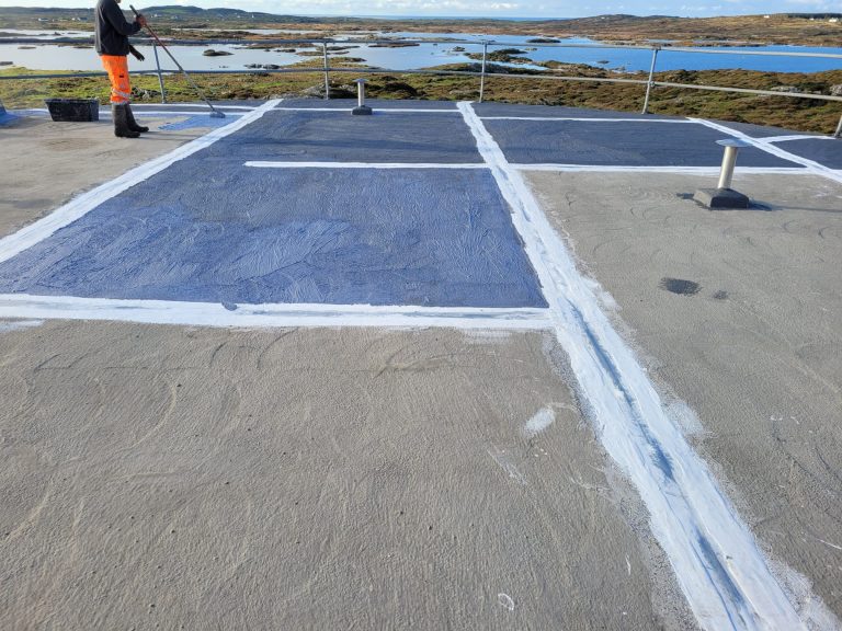 Person in orange pants applying a waterproof coating to a flat rooftop with a scenic view of water and rocky islands in the background.