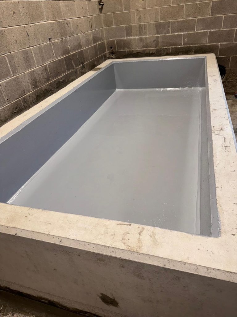 Concrete structure coated with a smooth gray waterproofing layer inside a tented construction area, featuring a blue folder resting on the right edge and two black pipes embedded in the dirt beside it.
