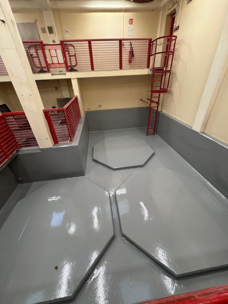 Interior view of an industrial or utility space with a shiny gray floor featuring three raised octagonal platforms, surrounded by walls painted cream and gray. The area is enclosed by red metal railings and includes a red vertical ladder with a safety cage attached to the wall. The space appears clean and well-maintained.
