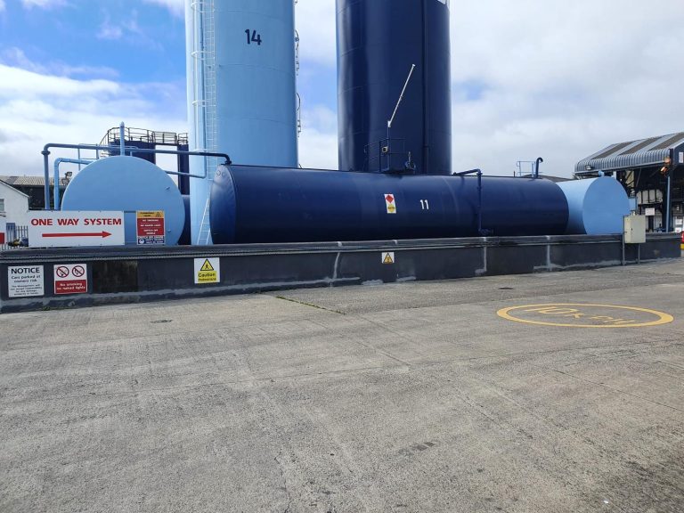 Large industrial storage tanks painted in blue at an outdoor facility, with safety signs including "One Way System," "Caution," and "Notice" visible along the perimeter.