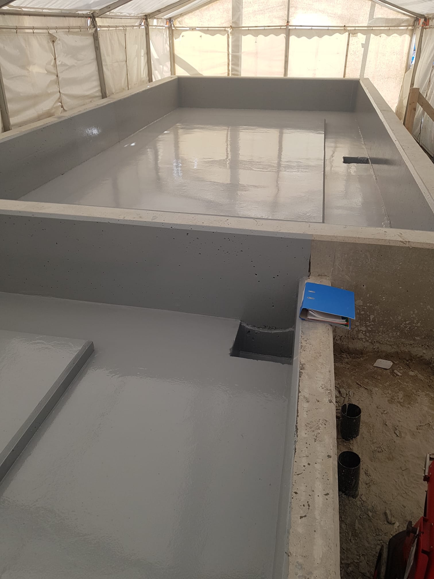 Concrete structure coated with a smooth gray waterproofing layer inside a tented construction area, featuring a blue folder resting on the right edge and two black pipes embedded in the dirt beside it.