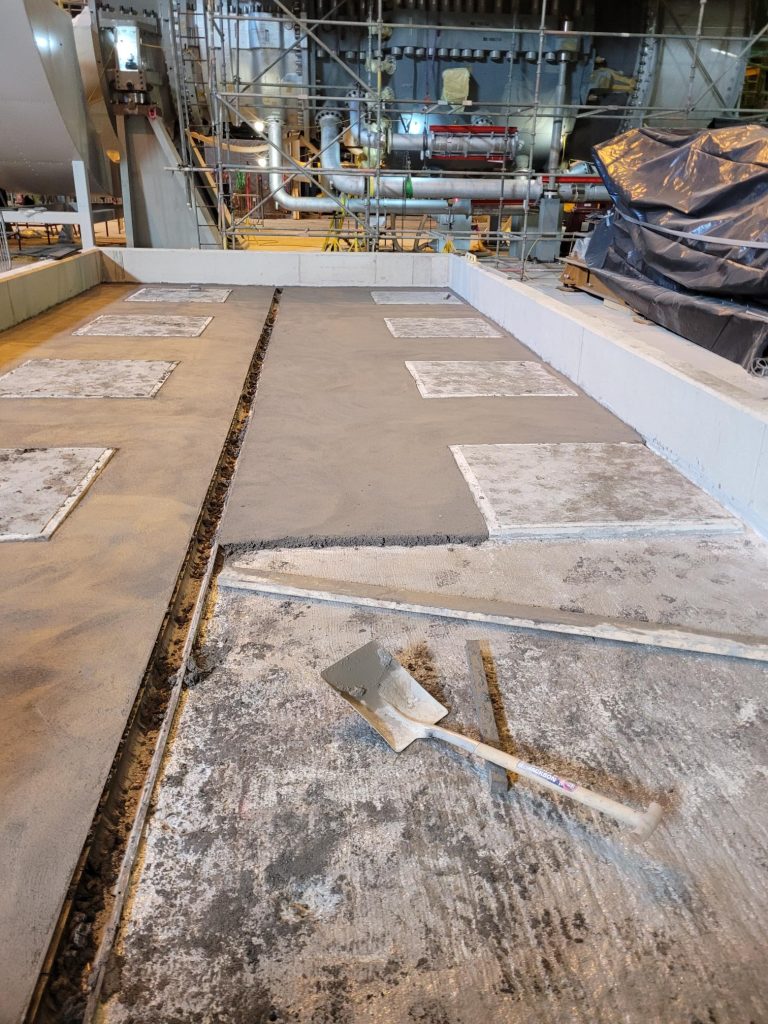 Concrete floor being repaired or resurfaced with a trowel lying on the ground, surrounded by industrial pipes and scaffolding in the background.
