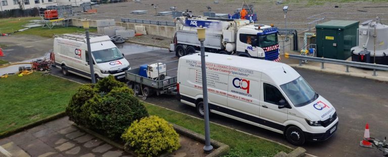 Two white vans with "Adrian Queally Ltd" logos parked side by side on a paved area, with one van towing a small trailer carrying equipment. In the background, a large white truck with "AQS" branding is parked near an industrial area with utility installations and a worker in an orange safety suit. Green shrubbery and a small grassy area are visible in the foreground.