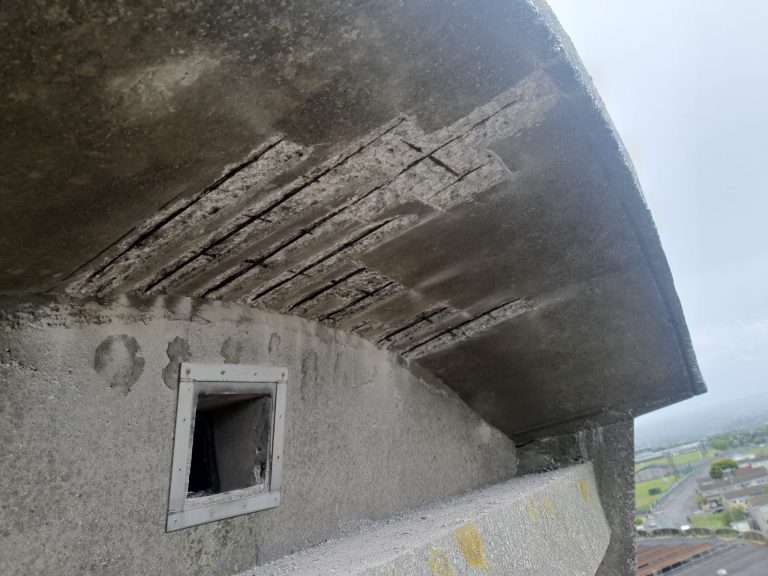 Close-up of a weathered concrete structure with visible cracks and exposed rebar on the underside of a projecting ledge, and a small rectangular window frame below. The background shows a cloudy sky and a distant urban landscape.