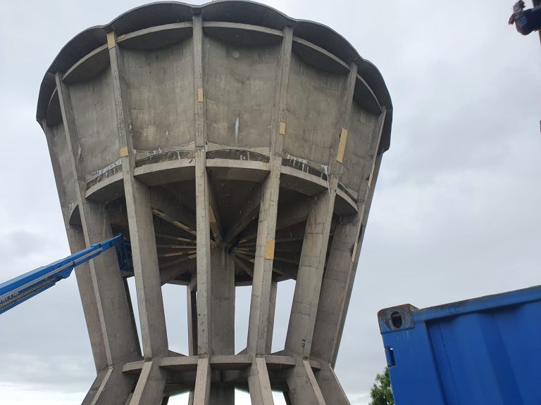 A large concrete water tower with a blue Genie hydraulic lift extended to the top, where two workers are performing maintenance. The area is fenced off with warning signs reading "Danger Men at Work" and "Danger Keep Out," under a partly cloudy sky.