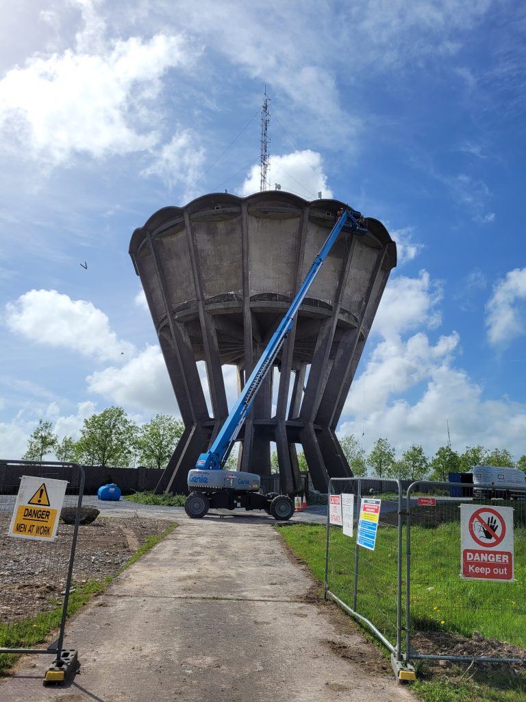 A large concrete water tower with a blue Genie hydraulic lift extended to the top, where two workers are performing maintenance. The area is fenced off with warning signs reading "Danger Men at Work" and "Danger Keep Out," under a partly cloudy sky.
