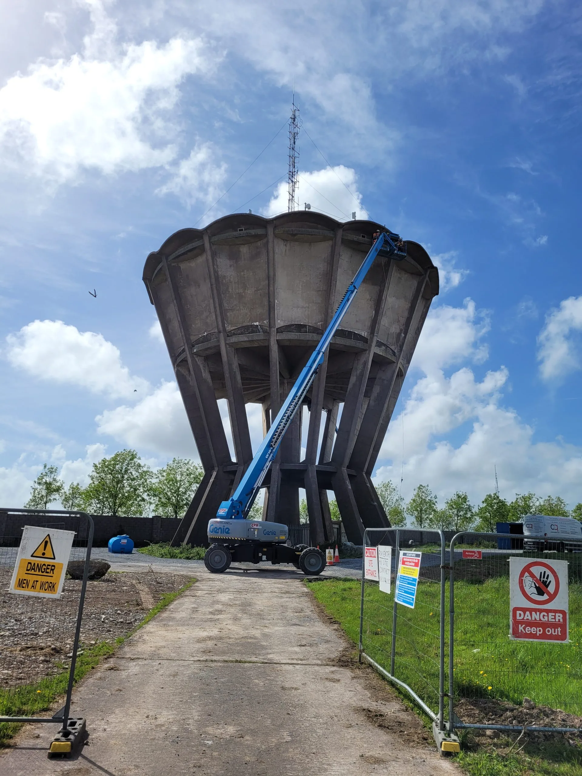 A large concrete water tower with a blue Genie hydraulic lift extended to the top, where two workers are performing maintenance. The area is fenced off with warning signs reading "Danger Men at Work" and "Danger Keep Out," under a partly cloudy sky.