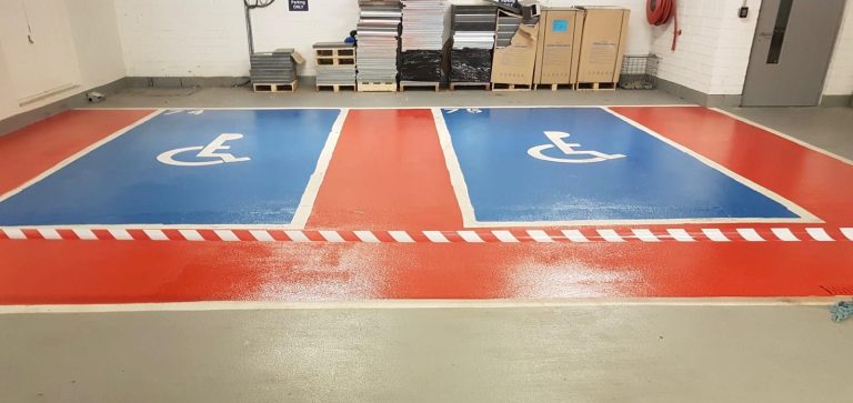 Two adjacent blue handicapped parking spaces with white wheelchair symbols, separated and bordered by red and white striped lines, inside an indoor area with stacks of boxes and pallets against the back wall.