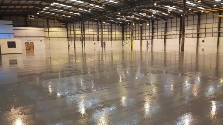 Large empty industrial warehouse with polished concrete floor and high ceiling with exposed beams and fluorescent lights.