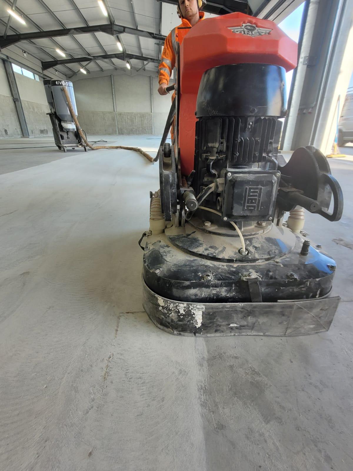 Worker wearing orange safety gear operates a floor grinding machine in a large industrial warehouse with concrete walls and a high ceiling.