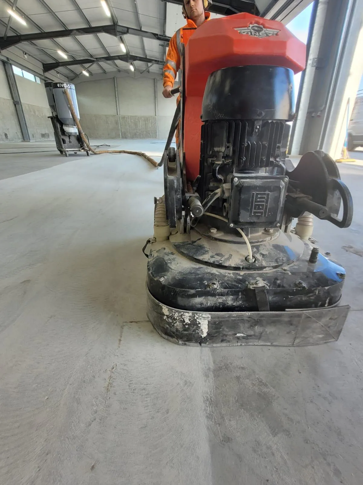 Worker wearing orange safety gear operates a floor grinding machine in a large industrial warehouse with concrete walls and a high ceiling.