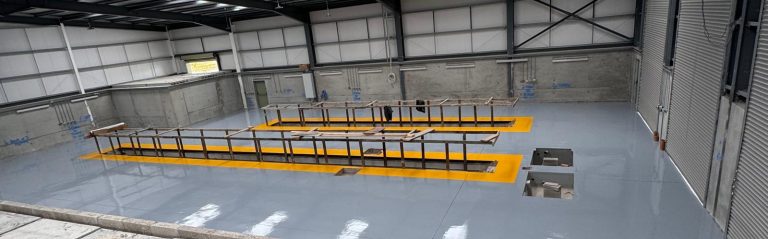 Empty industrial workshop with a clean, polished gray floor, yellow safety markings around two rectangular pits, and metal frames placed on top; concrete walls and large roller doors are visible.