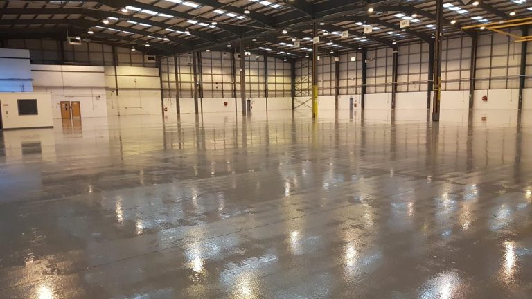 Large empty industrial warehouse with polished concrete floor and high ceiling with exposed beams and fluorescent lights.