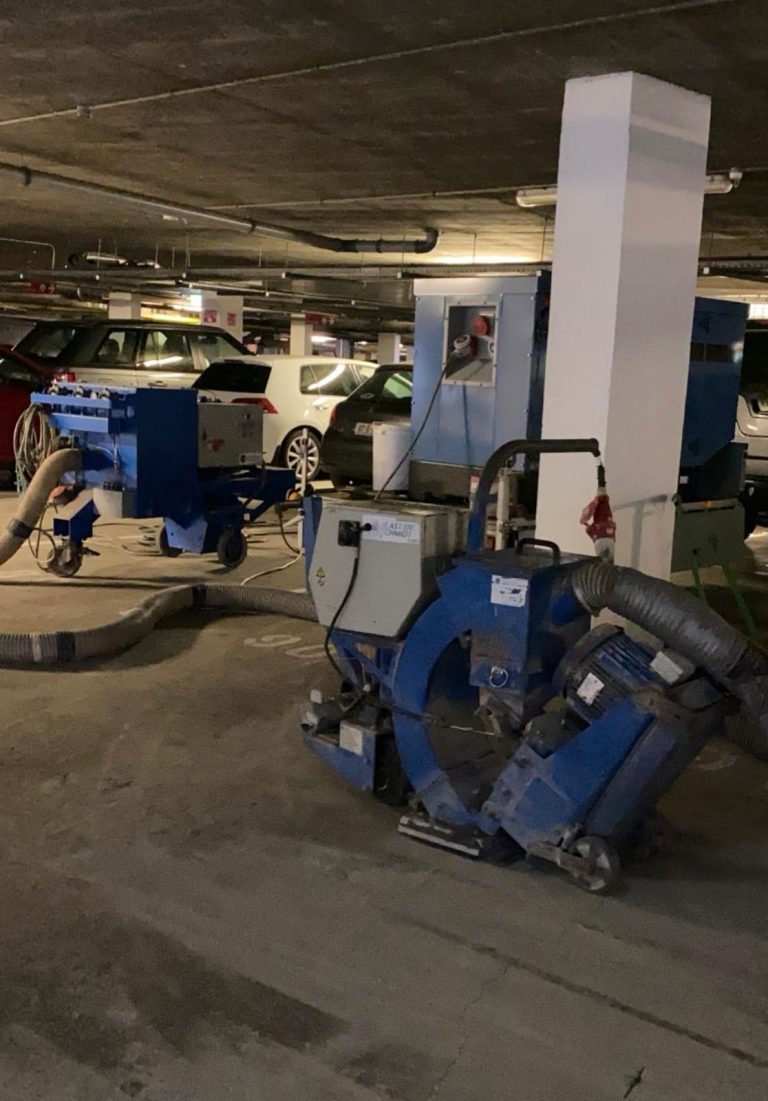 Industrial floor grinding machines with hoses attached are positioned in a dimly lit underground parking garage with several parked cars in the background.