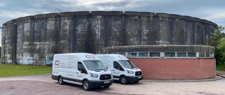 Two white vans parked in front of a large, weathered, circular concrete structure and a smaller brick building under a cloudy sky.