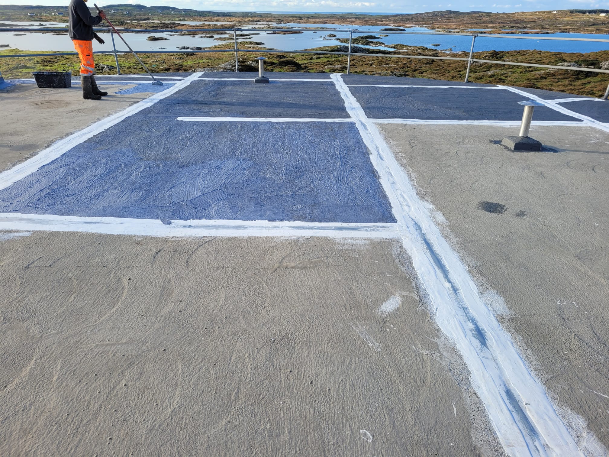 Person wearing orange work pants and black boots applying a blue coating to a sectioned flat rooftop near a body of water with rocky terrain in the background.