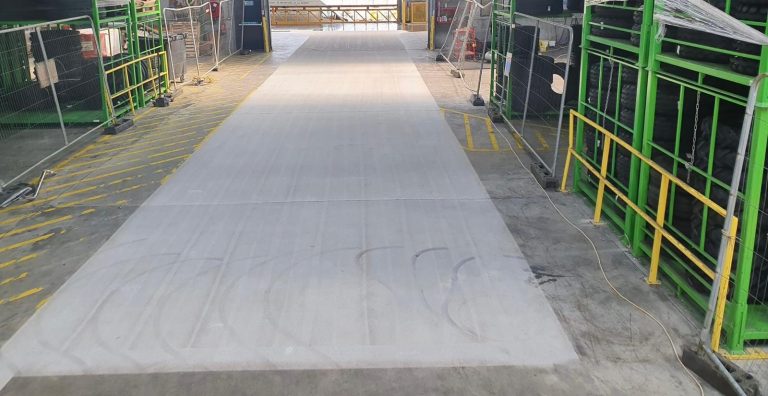 Industrial warehouse aisle with green metal racks holding black tires on both sides, yellow safety railings, and yellow diagonal floor markings. The aisle floor is mostly clean and clear, with some tire tread marks visible. The area is gated at the far end.