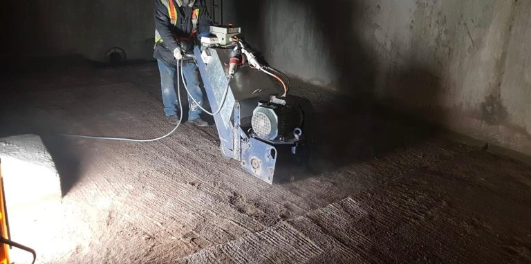 Worker using a heavy floor grinding machine to polish or level a rough concrete surface in a dimly lit indoor space with a bright work light illuminating the area.