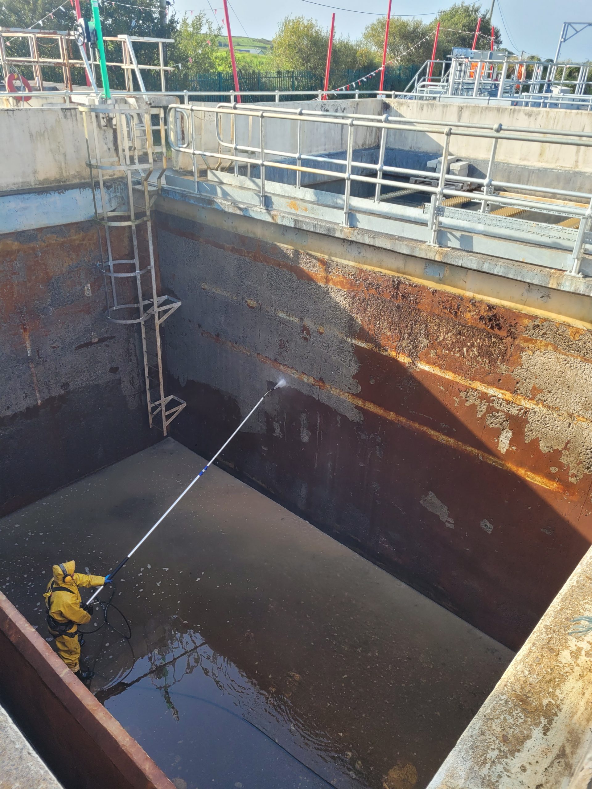 Person in a yellow protective suit pressure washing the interior wall of an industrial tank or large container with visible rust and grime.