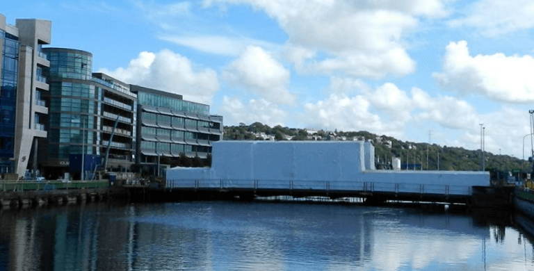 A river or canal with modern glass-office buildings on the left and a white, rectangular covered bridge stretching across the water under a partly cloudy blue sky.