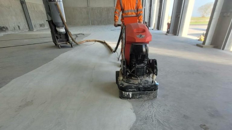A worker in orange safety gear is operating an industrial floor grinding machine on a large concrete surface inside a spacious building with multiple open garage-style doors.
