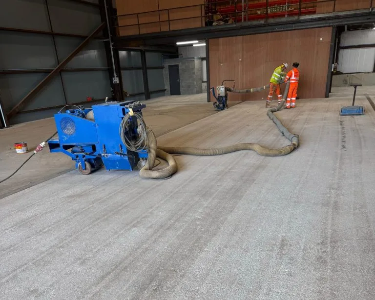 Two workers in high-visibility clothing operating a large blue industrial floor-cleaning or surface preparation machine inside a spacious warehouse or industrial building. The machine is connected by large hoses stretching across the concrete floor.
