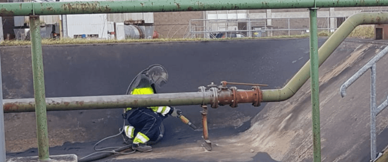 Worker in protective gear using a torch to perform maintenance or repair on a metal surface near industrial piping.