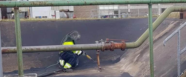 Worker in protective gear using a torch to perform maintenance or repair on a metal surface near industrial piping.